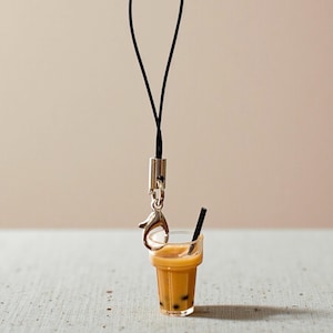 May include: A miniature charm of a bubble tea drink with a black straw. The drink is a light brown color with dark brown tapioca pearls at the bottom. The charm is attached to a black cord with a silver-colored clasp.