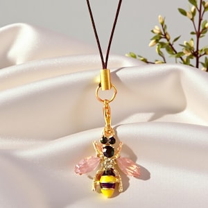 May include: A gold-colored bee pendant with pink and black crystal accents, and a yellow and black striped body. The pendant is attached to a brown cord with a gold clasp. The background is a white, silky fabric.