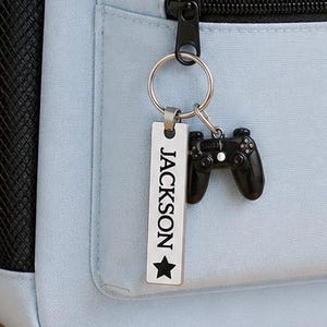 May include: A light blue backpack featuring a black mesh side pocket and a silver-toned metal keychain attached to the front zipper. The keychain includes a rectangular tag engraved with the name JACKSON and a star, alongside a black game controller charm.