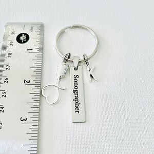 May include: Silver keychain with a heart charm, a small medical symbol charm, a letter K charm, and a rectangular tag that reads "Sonographer".