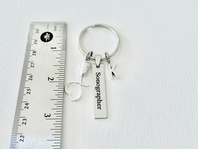 May include: Silver keychain with a heart charm, a small medical symbol charm, a letter K charm, and a rectangular tag that reads "Sonographer".