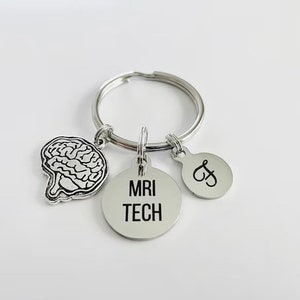 May include: A silver keychain with three charms. One charm is a brain outline, another is a round tag with "MRI TECH" in black, and the third is a small round tag with the letter "&" and the letter "J" in black. The keychain is on a white background.