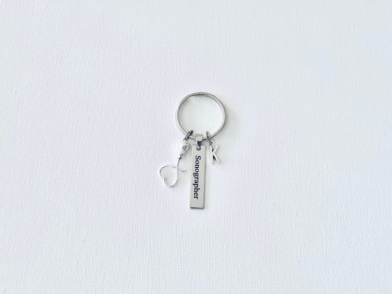 May include: Silver keychain with a heart charm, a letter K charm, and a rectangular tag that reads "Sonographer".