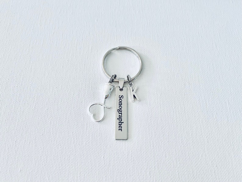 May include: Silver keychain with a heart charm, a letter K charm, and a rectangular tag that reads "Sonographer".