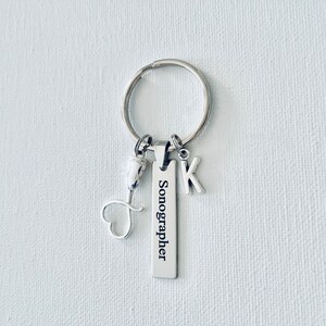 May include: Silver keychain with a charm that says "Sonographer" and a letter K charm. The keychain also has a small heart-shaped charm.