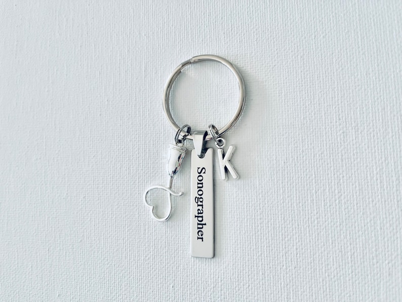 May include: Silver keychain with a charm that says "Sonographer" and a letter K charm. The keychain also has a small heart-shaped charm.