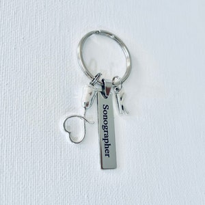 May include: Silver keychain with a heart charm, a small ultrasound probe charm, and a rectangular tag that reads "Sonographer".