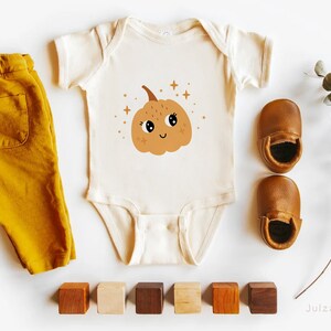 May include: A white baby onesie with a cute cartoon pumpkin with a smiling face and sparkly eyes. The onesie is surrounded by wooden blocks, brown leather baby shoes, and a sprig of eucalyptus.