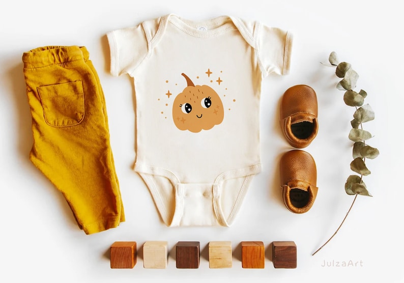 May include: A white baby onesie with a cute cartoon pumpkin with a smiling face and sparkly eyes. The onesie is surrounded by wooden blocks, brown leather baby shoes, and a sprig of eucalyptus.