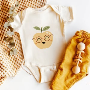 May include: A white baby onesie with a cute cartoon apple wearing glasses. The apple is yellow with a green stem and leaves. The onesie is lying on a brown knitted blanket with a pair of yellow pants and a wooden rattle.