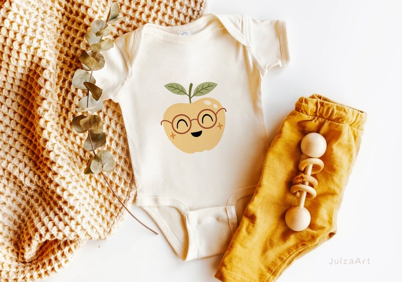 May include: A white baby onesie with a cute cartoon apple wearing glasses. The apple is yellow with a green stem and leaves. The onesie is lying on a brown knitted blanket with a pair of yellow pants and a wooden rattle.
