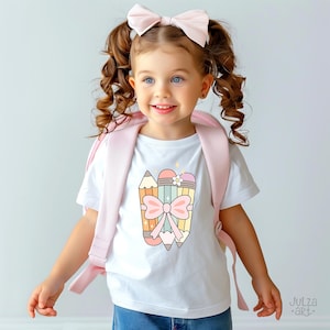 May include: A child wearing a white t-shirt featuring a colourful pencil graphic and a pink bow. The child also has a pink backpack and a matching bow in their hair. The image is from Julza Art.