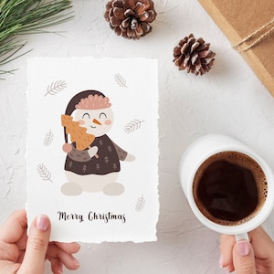 Pu&ograve; includere: Un biglietto bianco con un'illustrazione di un pupazzo di neve che tiene un piccolo albero arancione. Il pupazzo di neve indossa un cappello e un maglione marroni con un bordo bianco. Il biglietto ha il testo "Merry Christmas" scritto in corsivo nero.