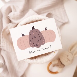 May include: A white card with a hand-drawn illustration of three pumpkins. The pumpkins are pink and brown with black crescent moons and dots. The text "Hello autumn!" is written in black below the pumpkins.