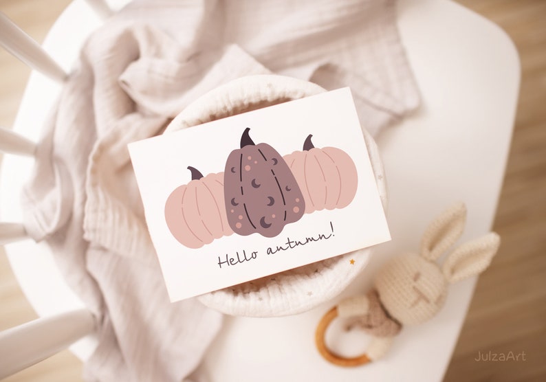 May include: A white card with a hand-drawn illustration of three pumpkins. The pumpkins are pink and brown with black crescent moons and dots. The text "Hello autumn!" is written in black below the pumpkins.