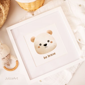 May include: A framed print featuring a watercolor illustration of a bear's face with the words "be brave" below. The artwork is in a white frame, and a crochet bunny rattle and a woven basket are nearby. The color palette is soft and neutral.