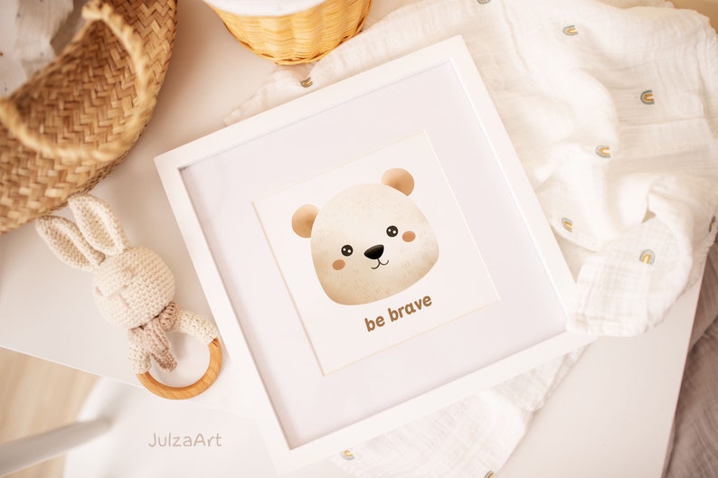 May include: A framed print featuring a watercolor illustration of a bear's face with the words "be brave" below. The artwork is in a white frame, and a crochet bunny rattle and a woven basket are nearby. The color palette is soft and neutral.