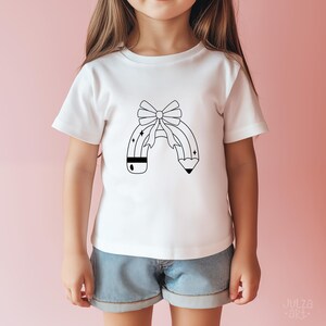 Op de afbeelding: Wit T-shirt met een zwart-wit potloodontwerp en een strik. Het potloodontwerp heeft een strik aan de bovenkant en sterren. Het shirt wordt door een kind gedragen.