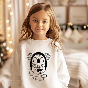 K&ouml;nnte beinhalten: Ein wei&szlig;er Pullover mit einer schwarz-wei&szlig;en Illustration eines Koalas, der einen Hut tr&auml;gt und eine Tasse h&auml;lt. Der Koala l&auml;chelt.