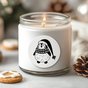 K&ouml;nnte beinhalten: Wei&szlig;e Kerze mit einer schwarz-wei&szlig;en Pinguin-Illustration auf dem Etikett. Der Pinguin tr&auml;gt einen Hut und einen Schal. Die Kerze befindet sich in einem Glasgef&auml;&szlig; mit einem wei&szlig;en Deckel.