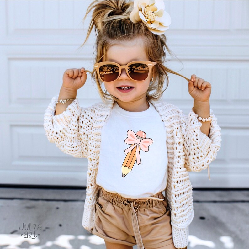 May include: A young child wearing a white t-shirt with a pencil graphic and a pink bow. The child is also wearing sunglasses, a cream-colored cardigan, and brown shorts. A flower hair accessory is visible.