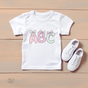 May include: White t-shirt with the letters "ABC" in pink and green, each adorned with a bow. The shirt is paired with white sneakers. The design is on a wooden surface.