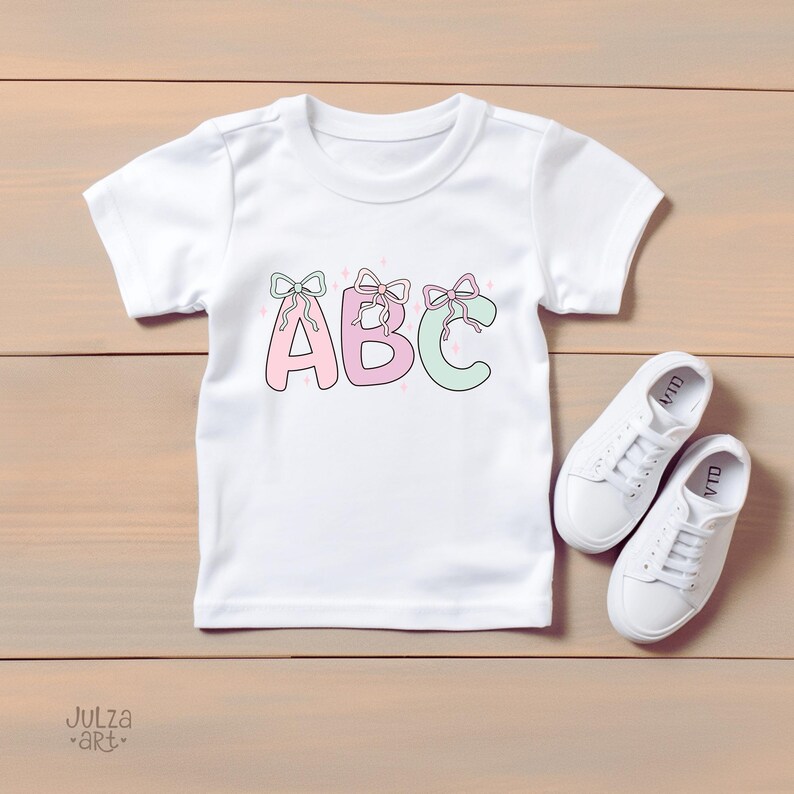 May include: White t-shirt with the letters "ABC" in pink and green, each adorned with a bow. The shirt is paired with white sneakers. The design is on a wooden surface.
