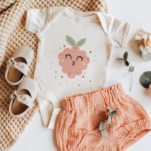 May include: A white baby onesie with a pink and green cartoon raspberry with a smiling face and winking eyes. The onesie is paired with a peach colored pair of baby bloomers.