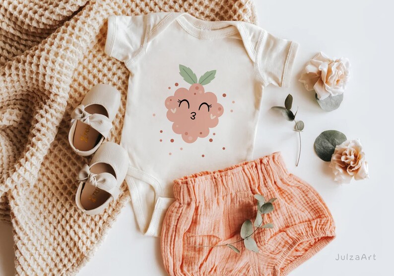 May include: A white baby onesie with a pink and green cartoon raspberry with a smiling face and winking eyes. The onesie is paired with a peach colored pair of baby bloomers.