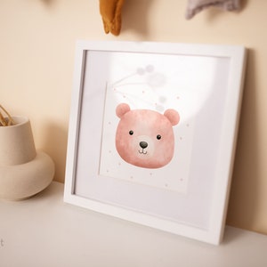 May include: A framed watercolor print featuring a pink bear face with a black nose and eyes. The bear is set against a white background with small pink dots. The frame is white, and the artwork is displayed on a white surface.
