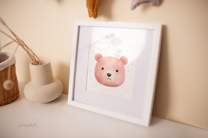 May include: A framed watercolor print featuring a pink bear face with a black nose and eyes. The bear is set against a white background with small pink dots. The frame is white, and the artwork is displayed on a white surface.