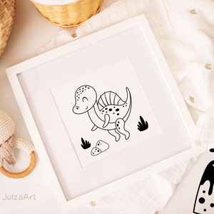 May include: Black and white line art print of a cartoon dinosaur with a spiky back, surrounded by small rocks and plants. The print is framed in a white frame and is sitting on a white surface.