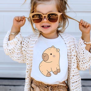 May include: A child wearing a white t-shirt featuring a cartoon capybara illustration in shades of tan and brown. The child is also wearing sunglasses, a cream-colored cardigan, and tan pants.