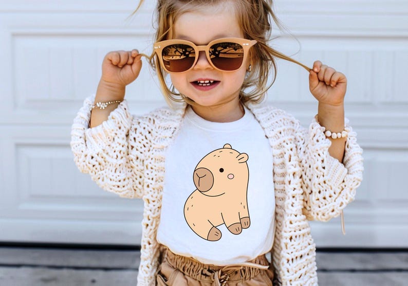 May include: A child wearing a white t-shirt featuring a cartoon capybara illustration in shades of tan and brown. The child is also wearing sunglasses, a cream-colored cardigan, and tan pants.