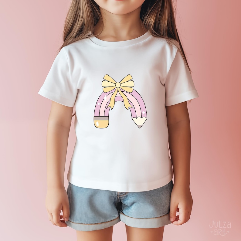 May include: White t-shirt with a pencil-shaped rainbow design and a yellow bow. The graphic uses pastel colours. The shirt is worn by a child. Denim shorts are also visible. The image has a pink background.