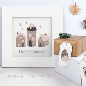 Puede incluir: Un marco blanco con una impresi&oacute;n de tres casas marrones con nieve en los techos y el texto "Happy Holidays". El marco est&aacute; sobre una superficie blanca con dos etiquetas de regalo, una con una manopla marr&oacute;n y la otra con una caja de regalo roja y blanca a rayas.