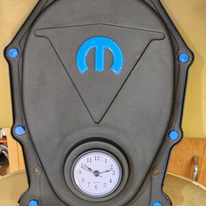 May include: A black clock with a blue Dodge logo and a white face. The clock is shaped like a timing chain cover and has blue bolts around the edge.
