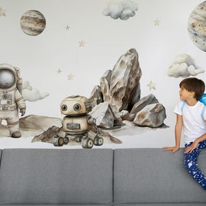 Può includere: Un bambino è seduto su un divano grigio di fronte a un muro decorato con adesivi murali a tema spaziale. Gli adesivi includono un robot, un astronauta, pianeti, stelle e rocce.