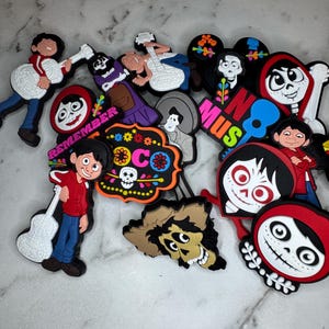 May include: A selection of colourful shoe charms featuring characters and imagery from the animated film "Coco." The charms include images of characters playing guitars, skulls, and the words "Remember" and "Music."
