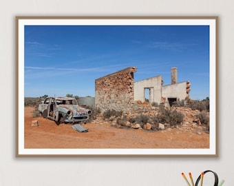 May include: A rusty car sits abandoned in front of a crumbling brick building in a desert landscape. The sky is blue and there are clouds in the distance.