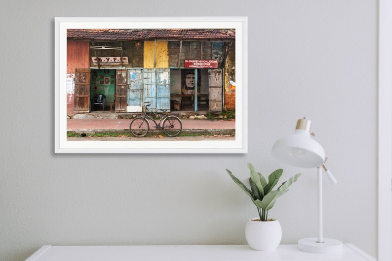 May include: A vintage bicycle is parked in front of a row of colorful, weathered storefronts in India. The storefronts have peeling paint and faded signs. The bicycle has a black frame, brown handlebars, and a brown seat.