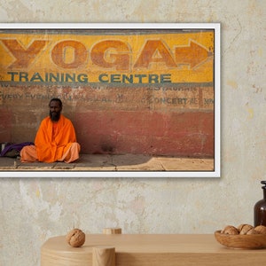 May include: A man in a bright orange robe sits in front of a wall with a faded sign that reads "Yoga Training Centre".