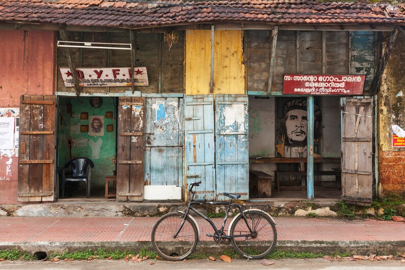 May include: A weathered wooden building with two open doorways. The building has a red, yellow, and blue paint scheme. A bicycle is parked in front of the building. The sign above the doorway on the left reads "D.Y.F.". The sign above the doorway on the right is in a foreign language.