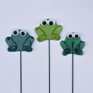 May include: Three glass frog garden stakes with white eyes and green bodies. The frogs are on metal stakes.