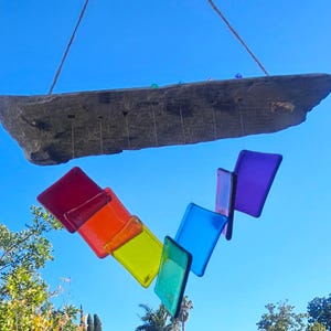 May include: A handcrafted wind chime with a natural stone top and colorful glass squares in a rainbow arrangement. The glass squares are red, orange, yellow, green, blue, and purple. The wind chime hangs from a natural fiber cord.
