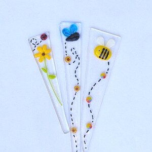 May include: Three clear glass plant stakes with colorful designs. One features a yellow flower and ladybug, another a blue and black bee, and the third a yellow and black bee. Each stake is approximately 6 inches long.