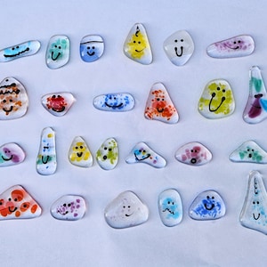 May include: A collection of 20 painted glass stones with various colors and designs. Each stone has a different smiley face drawn on it. The stones are arranged in a random pattern on a white background.