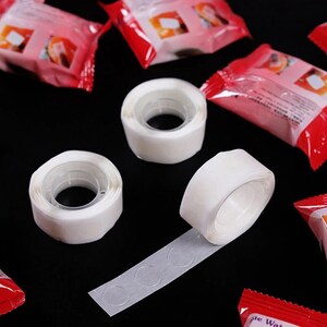 May include: Three white adhesive tape rolls with a clear adhesive backing. The tape is designed for attaching items to surfaces without damaging them. The tape is shown on a black background with red and white packaging.