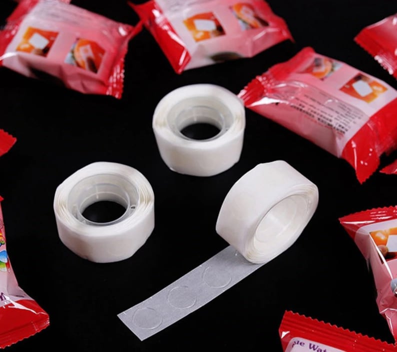 May include: Three white adhesive tape rolls with a clear adhesive backing. The tape is designed for attaching items to surfaces without damaging them. The tape is shown on a black background with red and white packaging.