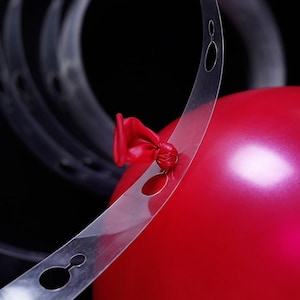 May include: Clear plastic balloon arch strip with holes for tying balloons. A red balloon is being tied to the strip.